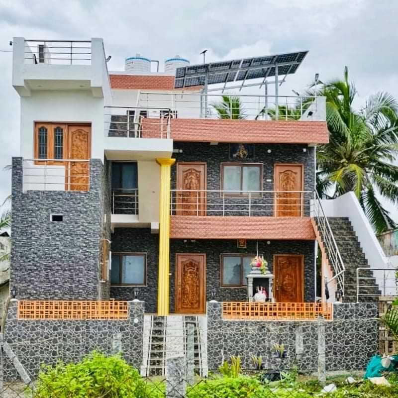 The house of SUHAMA. Multi-story building with textured stone facade, wooden doors, solar panels, and a small shrine at the entrance.